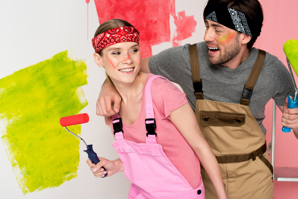 happy young couple in working overalls with painted faces holding paint rollers in front of painted wall  - Foto, Imagen