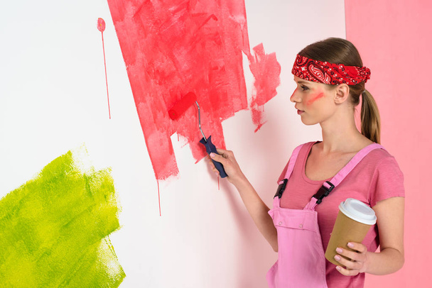 young woman with coffee cup painting wall in red by paint roller  - Foto, imagen
