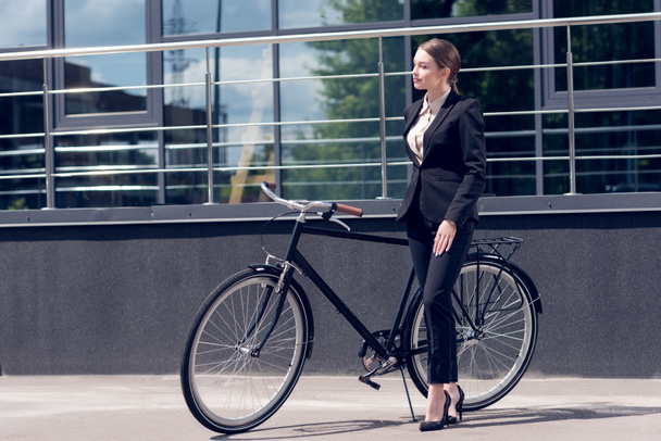 young businesswoman in suit and high heels standing near retro bicycle on street - Photo, image