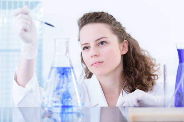 Chemist in a white coat holding a pipette and adding a blue substance to a laboratory flask, making a mixture - Fotografie, Obrázek