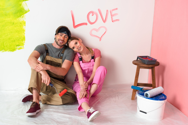 young couple in working overalls sitting near wall with lettering love and heart symbol  - Zdjęcie, obraz