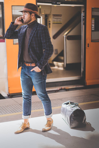 Atractivo y elegante joven hispano sosteniendo un teléfono inteligente en su mano y llamando mientras está de pie en la plataforma ferroviaria esperando el tren.
. - Foto, imagen