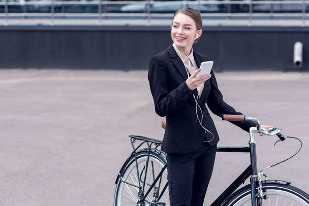 portrait of smiling businesswoman in earphones with smartphone standing near retro bicycle on street - Photo, Image