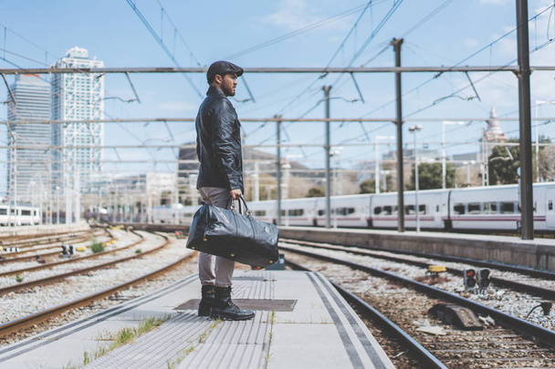 Attractive confident hispanic man having a backpack front of him with a clothes bag, walking beside a platform at train station. - Фото, зображення