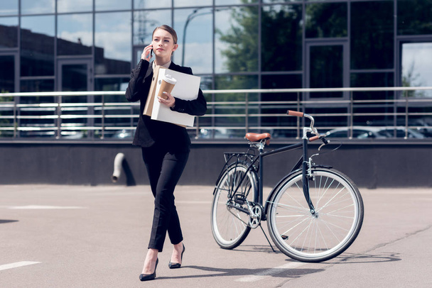 businesswoman with documents and coffee to go talking on smartphone while walking on street with bicycle parked behind - 写真・画像