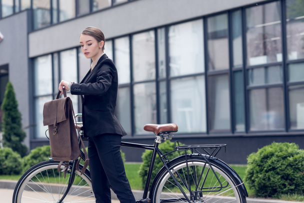 portrait of young businesswoman with briefcase and retro bicycle checking time on street - 写真・画像
