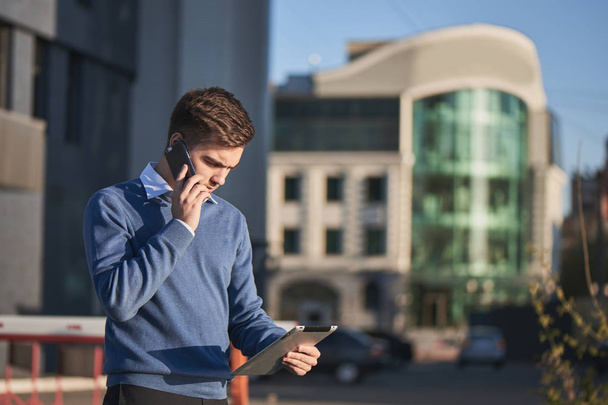 hombre de negocios guapo en jersey y camisa hablando por teléfono y mirando la tableta PC en la mano al aire libre
  - Foto, Imagen