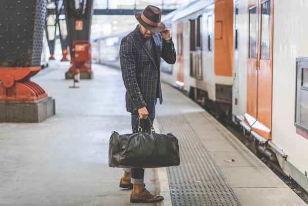 Attraente hipster ispanico sicuro di sé con borsa da viaggio con vestiti casual e cappello in attesa del treno sulla piattaforma ferroviaria
 - Foto, immagini
