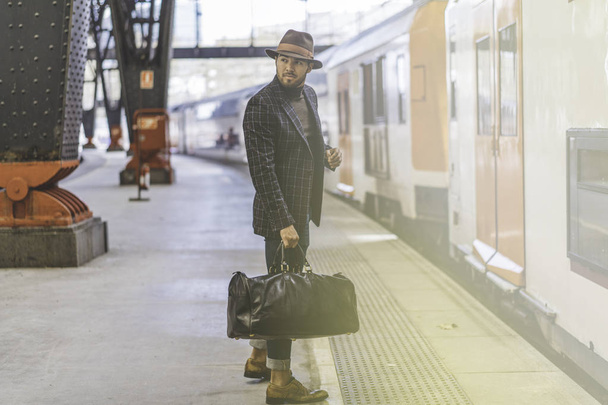 Guapo hipster hispano confiado con bolsa de viaje con ropa casual y sombrero esperando el tren en la plataforma ferroviaria
 - Foto, imagen