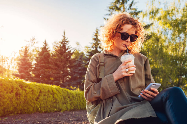 Jeune femme hippie aux cheveux roux bouclés buvant du café et utilisant un smartphone dans un parc d'été. Fille élégante se refroidissant à l'extérieur au coucher du soleil
 - Photo, image