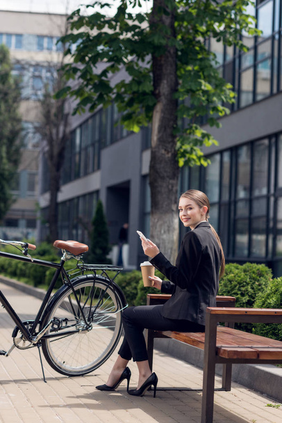 usměvavá mladá podnikatelka s smartphone a kávu jít opřenou o lavičku - Fotografie, Obrázek