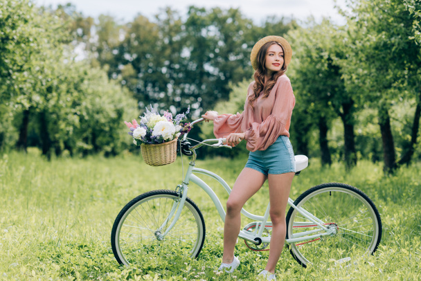 young beautiful woman standing near retro bicycle with wicker basket full of flowers in forest - Photo, Image