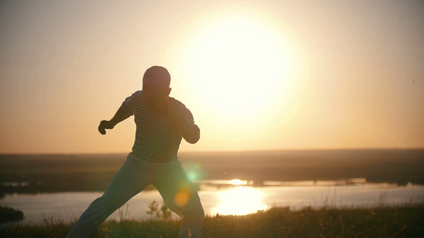 Боєць виконує бойові трюки з танцювальними елементами на пагорбі на літньому заході сонця
 - Кадри, відео