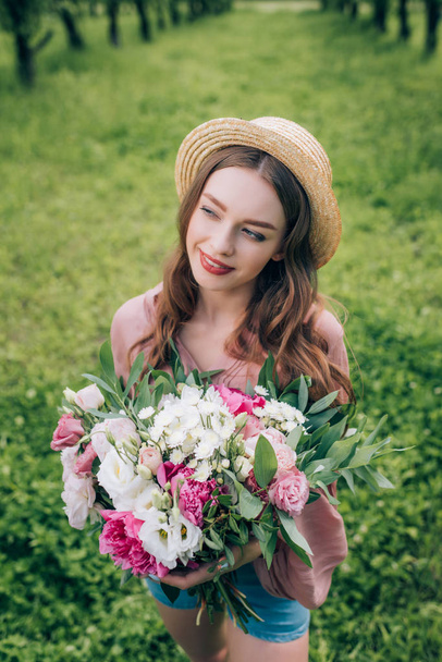 high angle view of beautiful smiling woman in hat with bouquet of flowers looking away in park - Foto, immagini