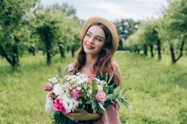 портрет красивой улыбающейся женщины в шляпе с букетом цветов, смотрящей в парк
 - Фото, изображение