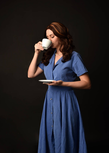 3/4 青いドレスを着て、紅茶のカップを持ってブルネットの女性の肖像画。黒のスタジオの背景にもたらされる. - 写真・画像