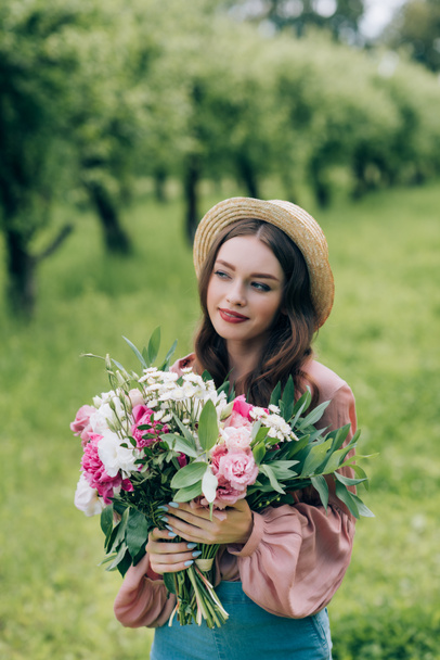 portrait of beautiful pensive woman in hat with bouquet of flowers in park - Фото, изображение
