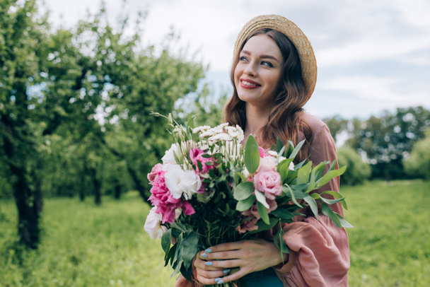 portrait of pretty young woman in hat with bouquet of flowers in park - Photo, Image