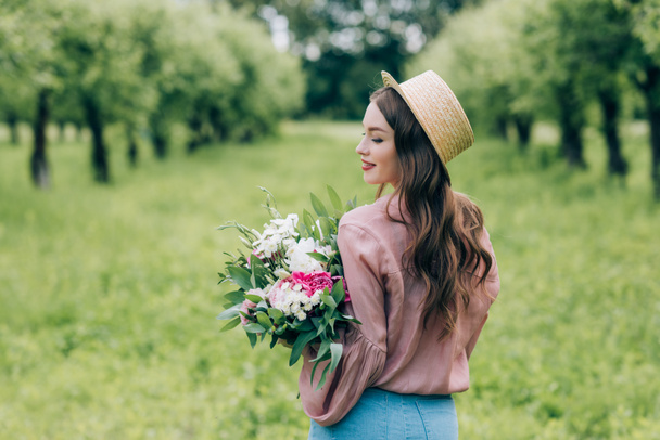 back view of smiling woman in hat with bouquet of flowers in hands standing in park - Photo, Image