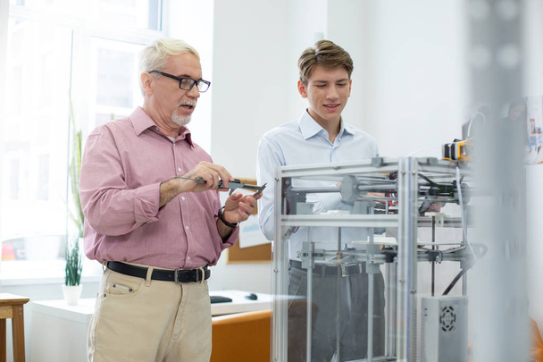 Senior engineer using caliper while printing models with intern - Foto, Bild