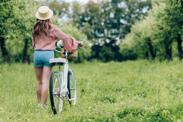 back view of young woman with retro bicycle with wicker basket full of flowers in forest - Foto, imagen