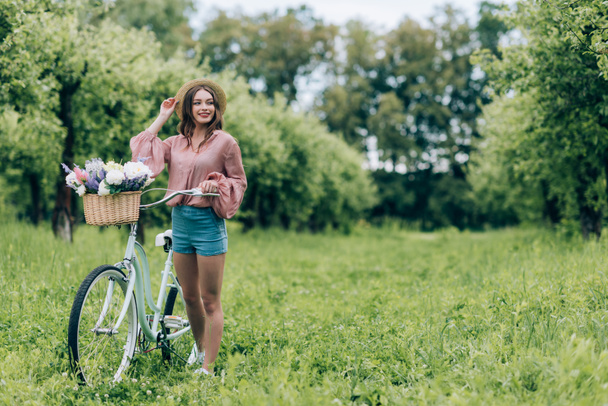 young pretty woman holding retro bicycle with wicker basket full of flowers in forest - Fotoğraf, Görsel