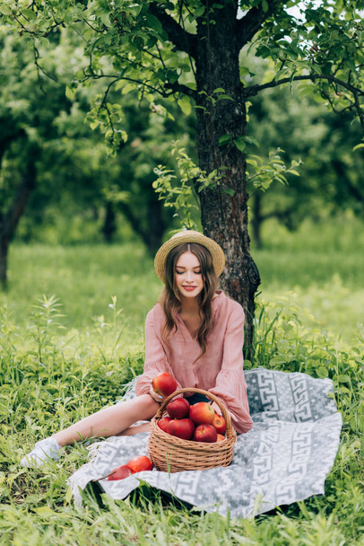 pretty young woman in hat resting on blanket with wicker basket with apples in park - Foto, immagini