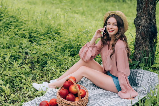 joven mujer sonriente hablando en el teléfono inteligente mientras descansa sobre una manta con canasta de mimbre llena de manzanas en el parque
 - Foto, Imagen
