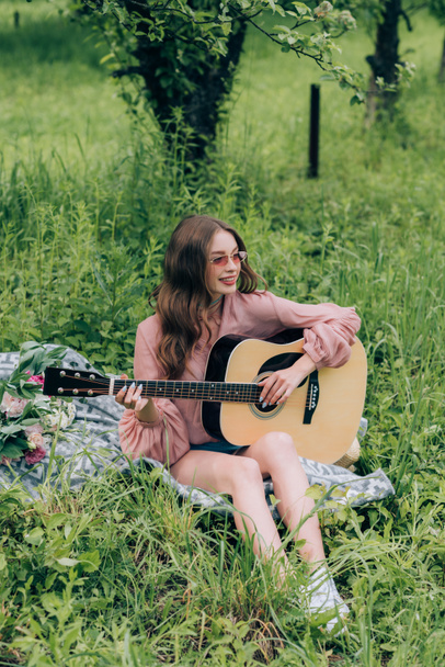 fiatal nő játszik akusztikus gitár, miközben pihen a csokor virágot parkban takaró - Fotó, kép