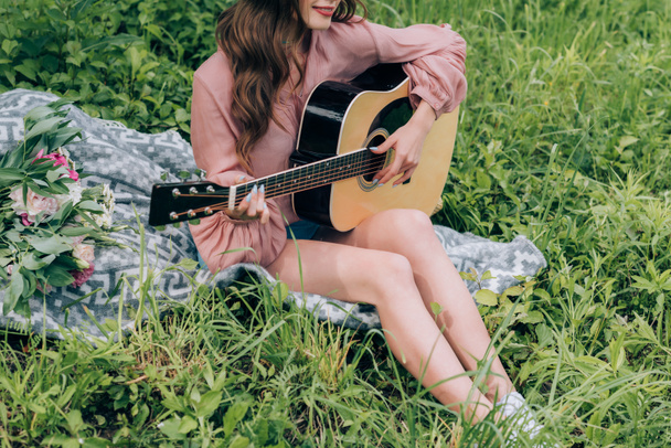 частковий вид на жінку, яка грає на акустичній гітарі, відпочиваючи на ковдрі з букетом квітів у парку
 - Фото, зображення