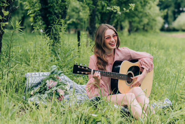 happy woman in sunglasses with acoustic guitar resting on blanket with bouquet of flowers in park - 写真・画像