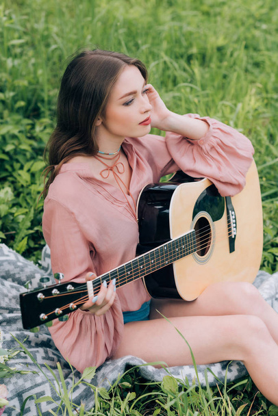 young pensive woman with acoustic guitar resting on blanket with bouquet of flowers in park - Foto, afbeelding