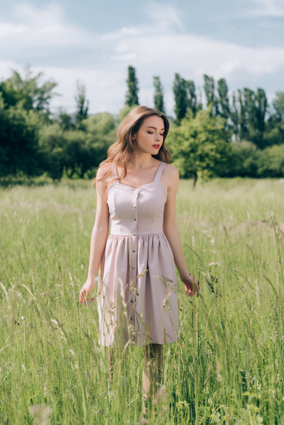 joven mujer pensativa en vestido elegante con el pelo largo caminando en el prado solo
 - Foto, Imagen