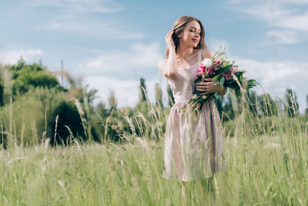 portrait of beautiful young woman holding bouquet of flowers while standing in field alone - Photo, Image