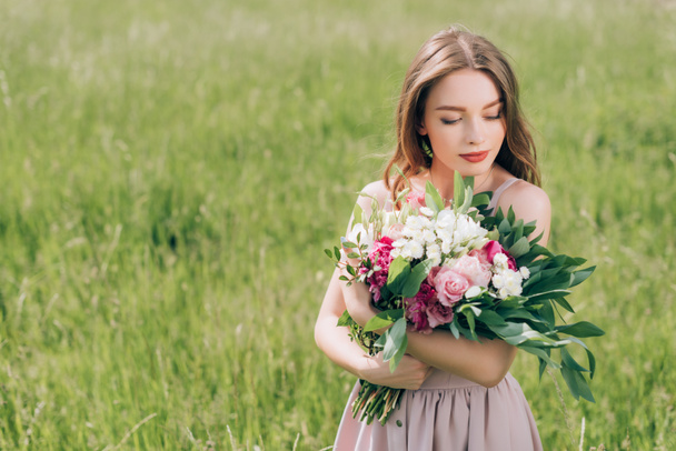 portrait of beautiful pensive woman holding bouquet of flowers while standing in field alone - Фото, изображение