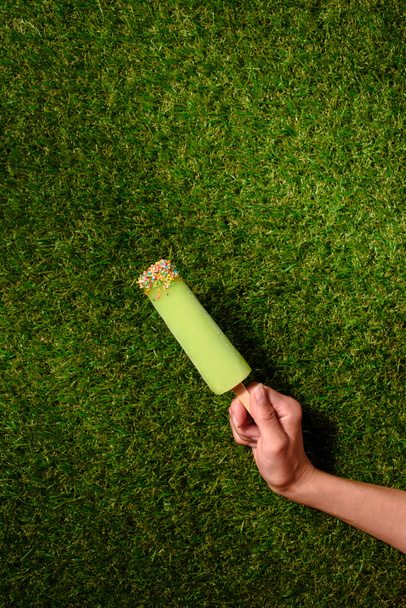Рука тримає фруктовий лід поп на фоні зеленої трави
 - Фото, зображення