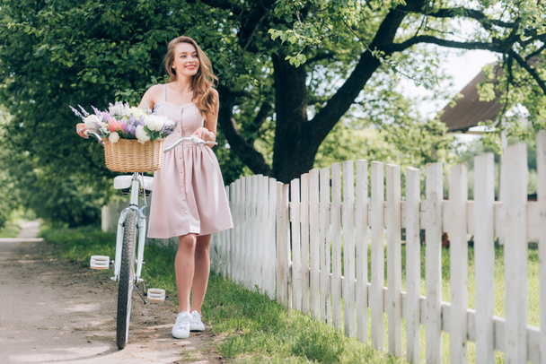 hermosa mujer sonriente en vestido con bicicleta retro con canasta de mimbre llena de flores en el campo
 - Foto, Imagen