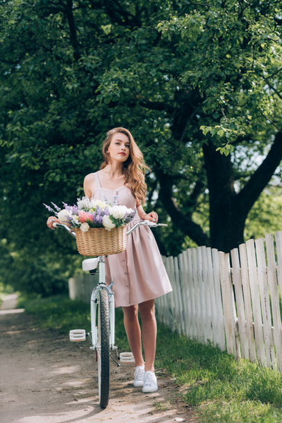 bella donna pensierosa in abito con bicicletta retrò con cesto di vimini pieno di fiori in campagna
 - Foto, immagini