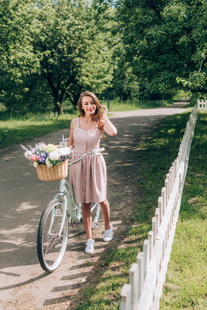 young beautiful woman in dress with retro bicycle with wicker basket full of flowers at countryside - Фото, зображення