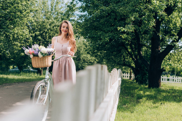 young beautiful woman in dress with retro bicycle with wicker basket full of flowers at countryside - Fotografie, Obrázek