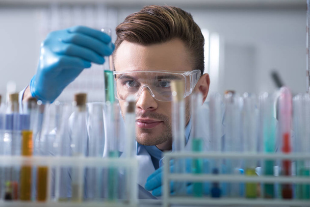 Gripping substance. Young interested beardful scientist in a glasses and rubber gloves working in the lab keeping and overlooking test tube. - Photo, image