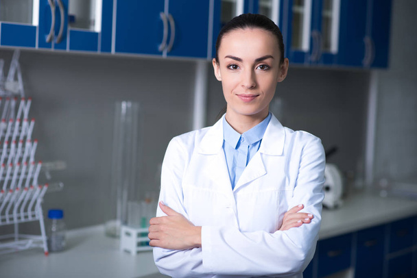 Best expert. Pretty young beautiful discoverer in a uniform standing in the laboratory smiling and holding crossed her hands. - Fotografie, Obrázek