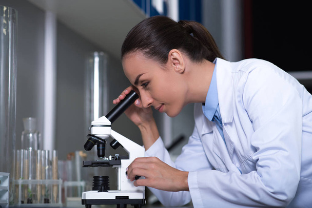 Microbiologists reality. Adult cute reasonable scientist in a uniform standing in the lab working with the microscope and looking in the scope. - Zdjęcie, obraz