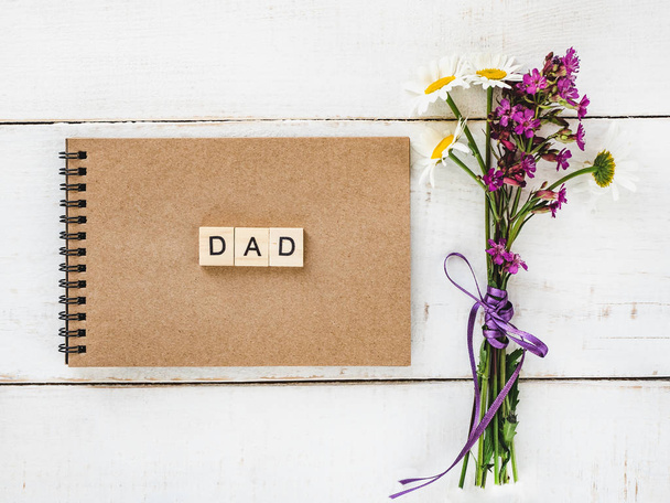 Notepad with wooden letters of the alphabet and the word DAD, a bouquet wildflowers on a white, vintage, wooden table. Top view, close-up. Congratulations for loved ones and relatives - 写真・画像