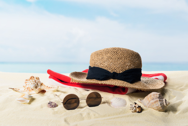 Strooien hoed en zonnebril met schelpen in het zand op de blauwe hemelachtergrond - Foto, afbeelding