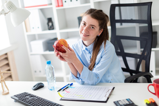 Genç bir kız ofiste bir masada oturur ve bir elma elinde tutan. - Fotoğraf, Görsel