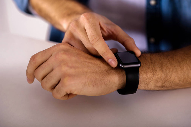 Modern user. Close up of pleasant man using his smart wrist watch while standing near table - Foto, afbeelding