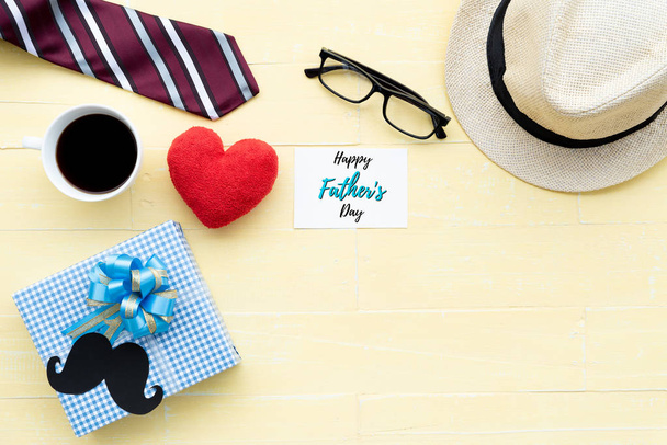 Happy fathers day concept. Red tie, glasses, hat, mustache, gift box with Love DAD text on black heart tag on bright yellow pastel wooden table background. - Photo, Image