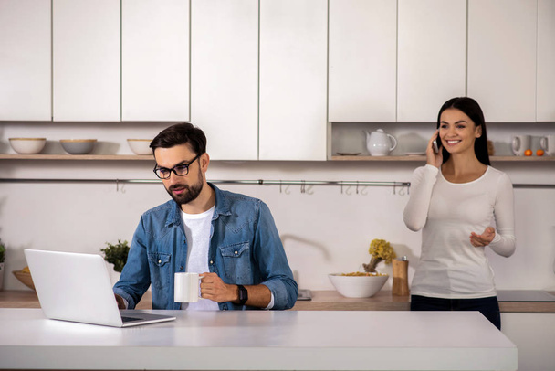 Ranní tradice. Pozitivní vousatý muž pomocí přenosného počítače a jeho přítelkyně mluví o telefonu v kuchyni - Fotografie, Obrázek