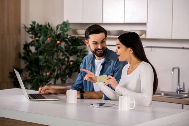 Positive emotions. Cheerful yougn couple sititng in the kitchen while using the online banking service - Фото, изображение